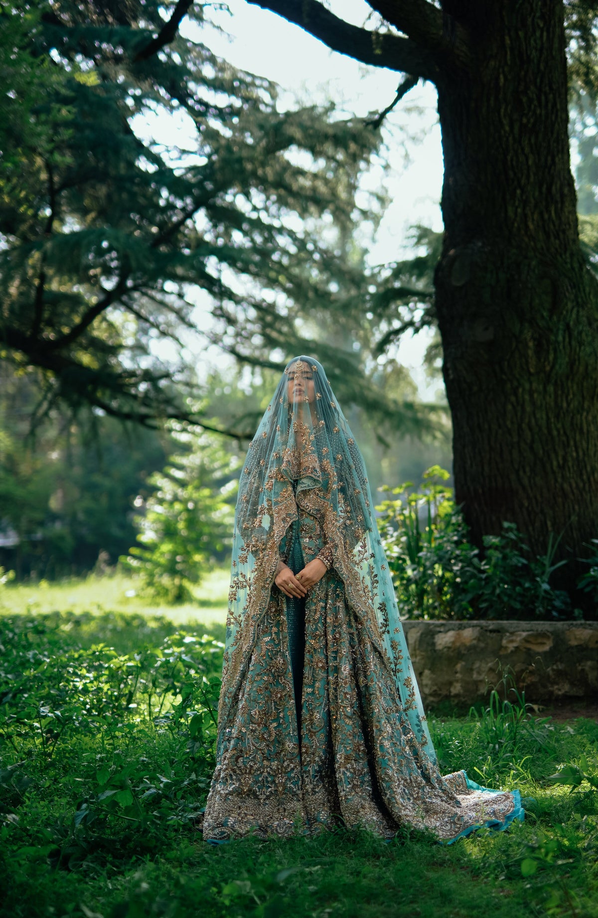 A woman stands outdoors in a lush garden wearing an ornate, floor-length teal and gold embroidered gown with a sheer matching veil.