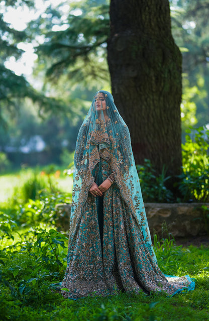 A woman stands outdoors in a lush garden wearing an ornate teal and gold embroidered bridal gown with a matching sheer veil and long train.