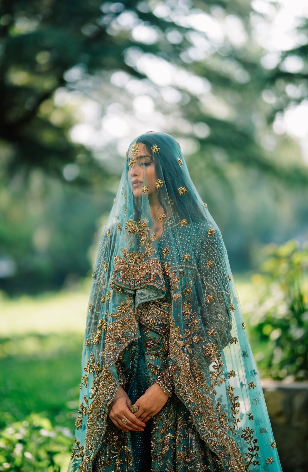 A woman wearing an ornate teal and gold embroidered traditional outfit with a sheer teal veil, standing outdoors in a garden setting with blurred green foliage in the background.