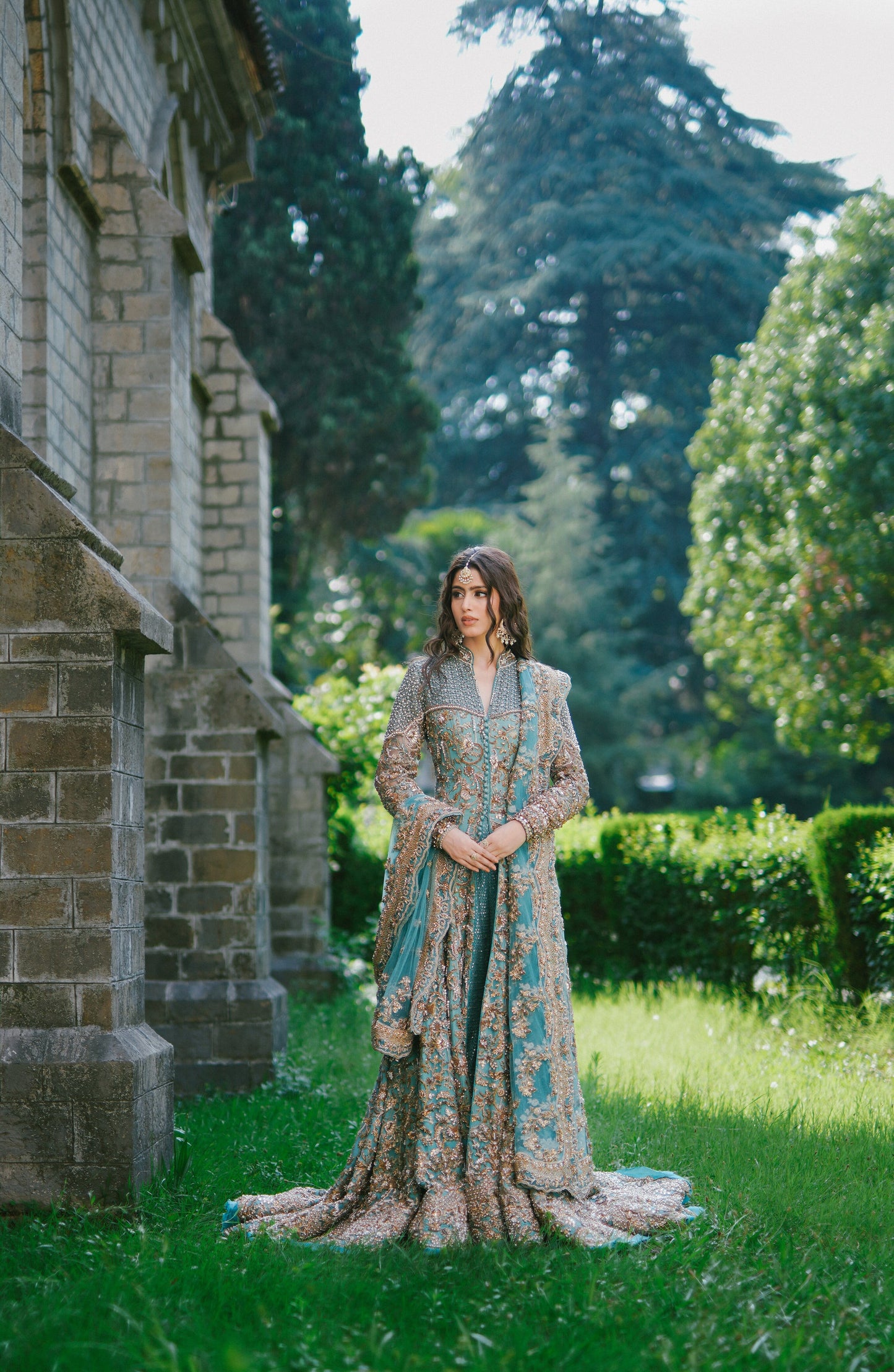 A woman stands outdoors on a grassy lawn beside a stone building, wearing a long, heavily embroidered gown in muted teal, beige, and gold tones. Trees and hedges are visible in the background.