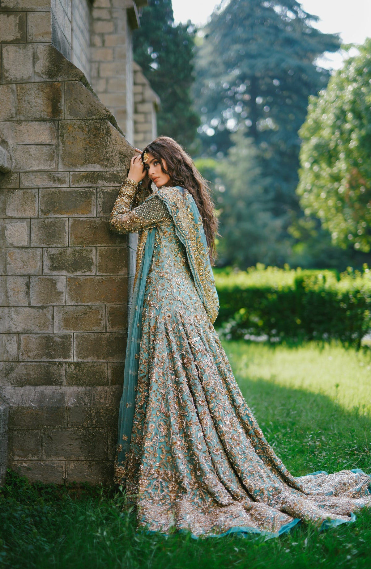 A woman wearing a long, heavily embroidered traditional dress leans against a stone wall in an outdoor garden setting. The dress features a complex floral and paisley pattern in muted teal, beige, and brown tones, with a matching dupatta draped over her shoulder.