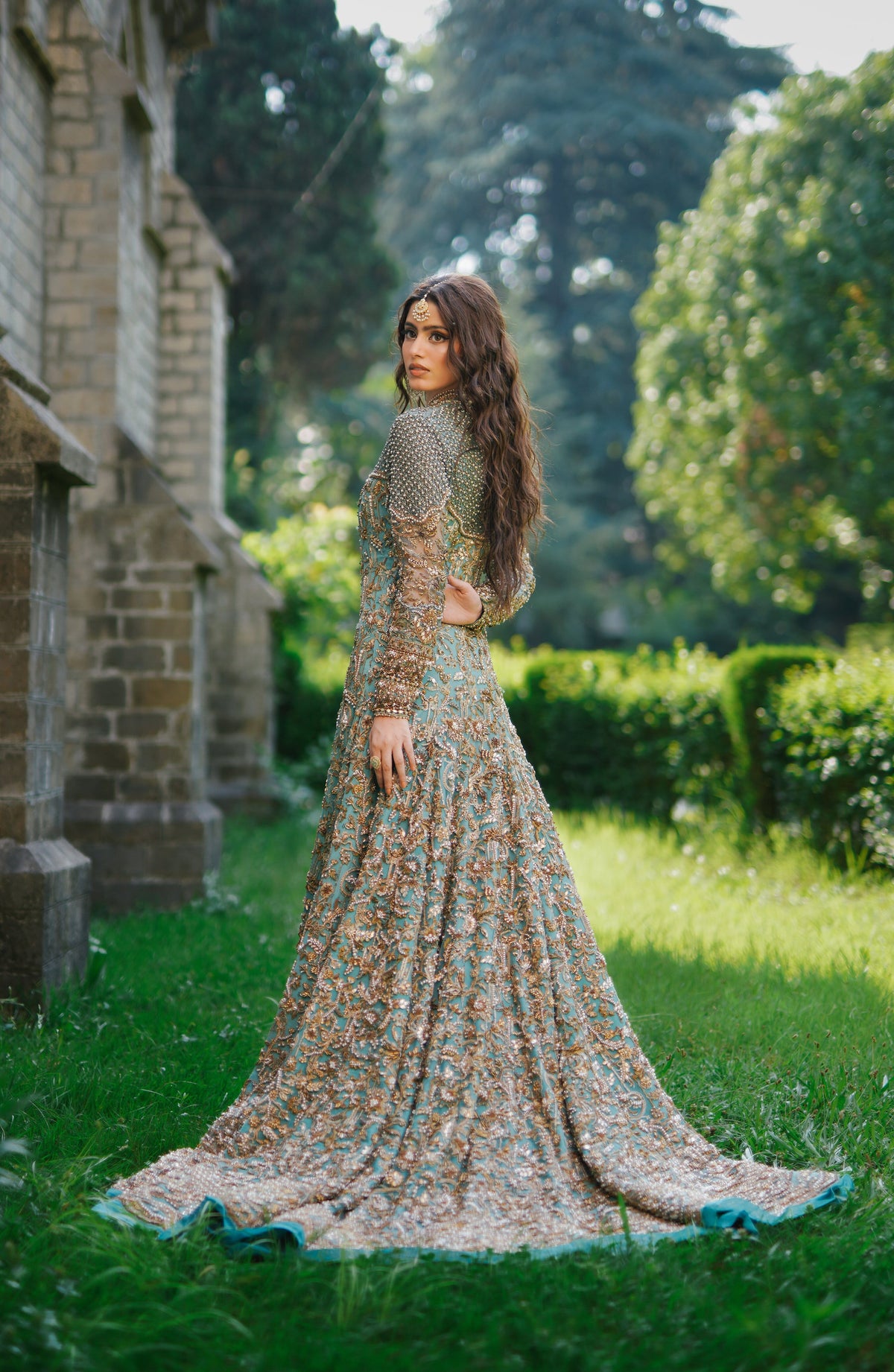 A woman stands outdoors in a lush garden next to a stone building, wearing a long-sleeved, floor-length embellished gown with a multicolor floral pattern and a turquoise border.
