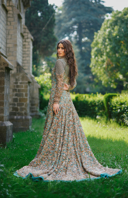 A woman stands outdoors in a lush garden next to a stone building, wearing a long-sleeved, floor-length embellished gown with a multicolor floral pattern and a turquoise border.
