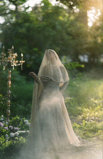A woman in a long sheer white bridal veil and fitted lace wedding gown stands outdoors in a garden at golden hour, with soft sunlight, blurred greenery, flowers, and a decorative candelabra visible.