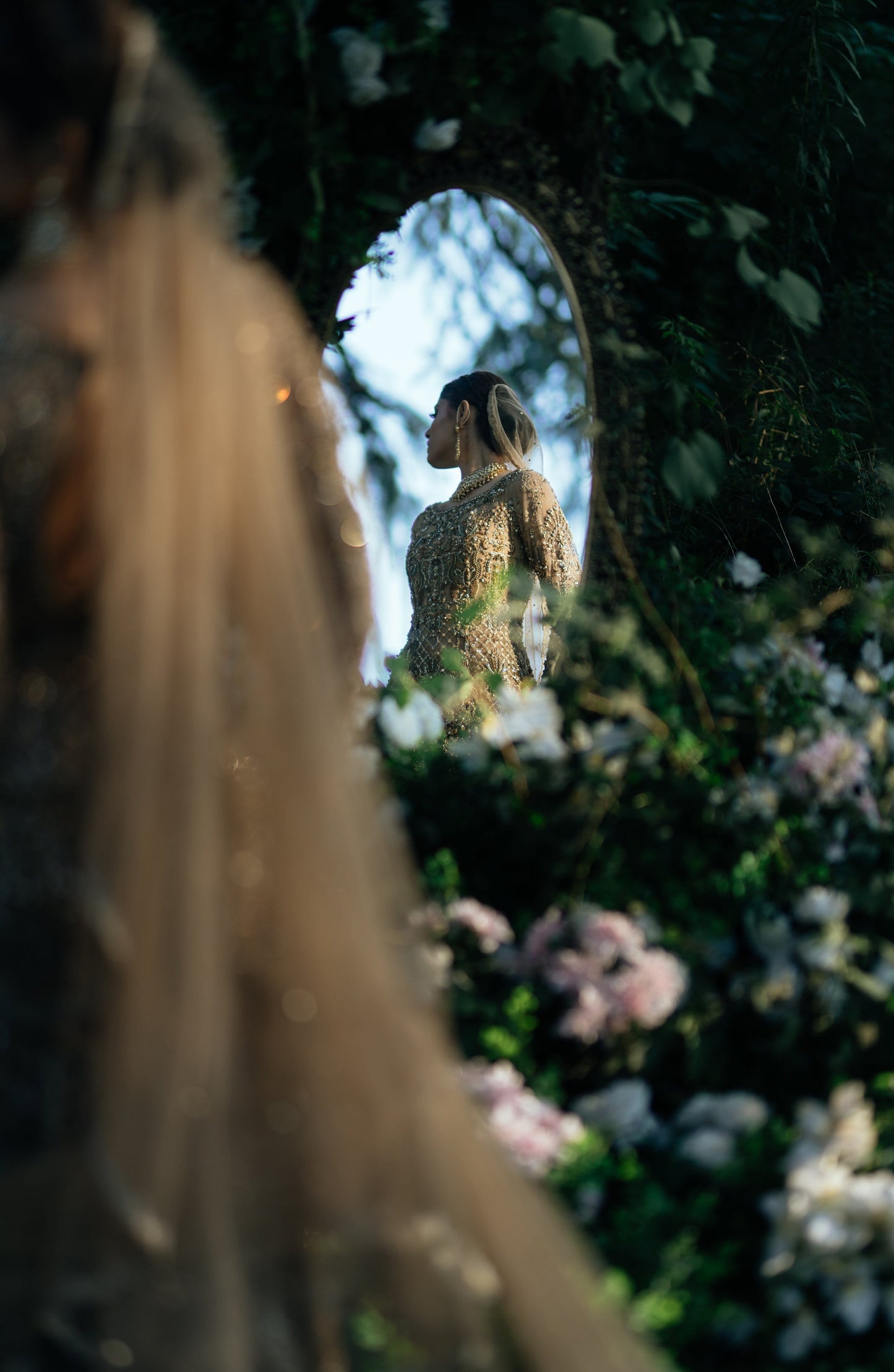 A woman in a textured beige dress stands in front of an oval mirror framed by lush green foliage and soft pink flowers. The foreground is heavily blurred, drawing focus to the subject in the mirror reflection.