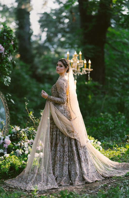 A woman wearing an ornate long beige and brown embroidered gown with a sheer draped overlay stands outdoors in a garden setting with trees, flowers, and a candelabra in the background.