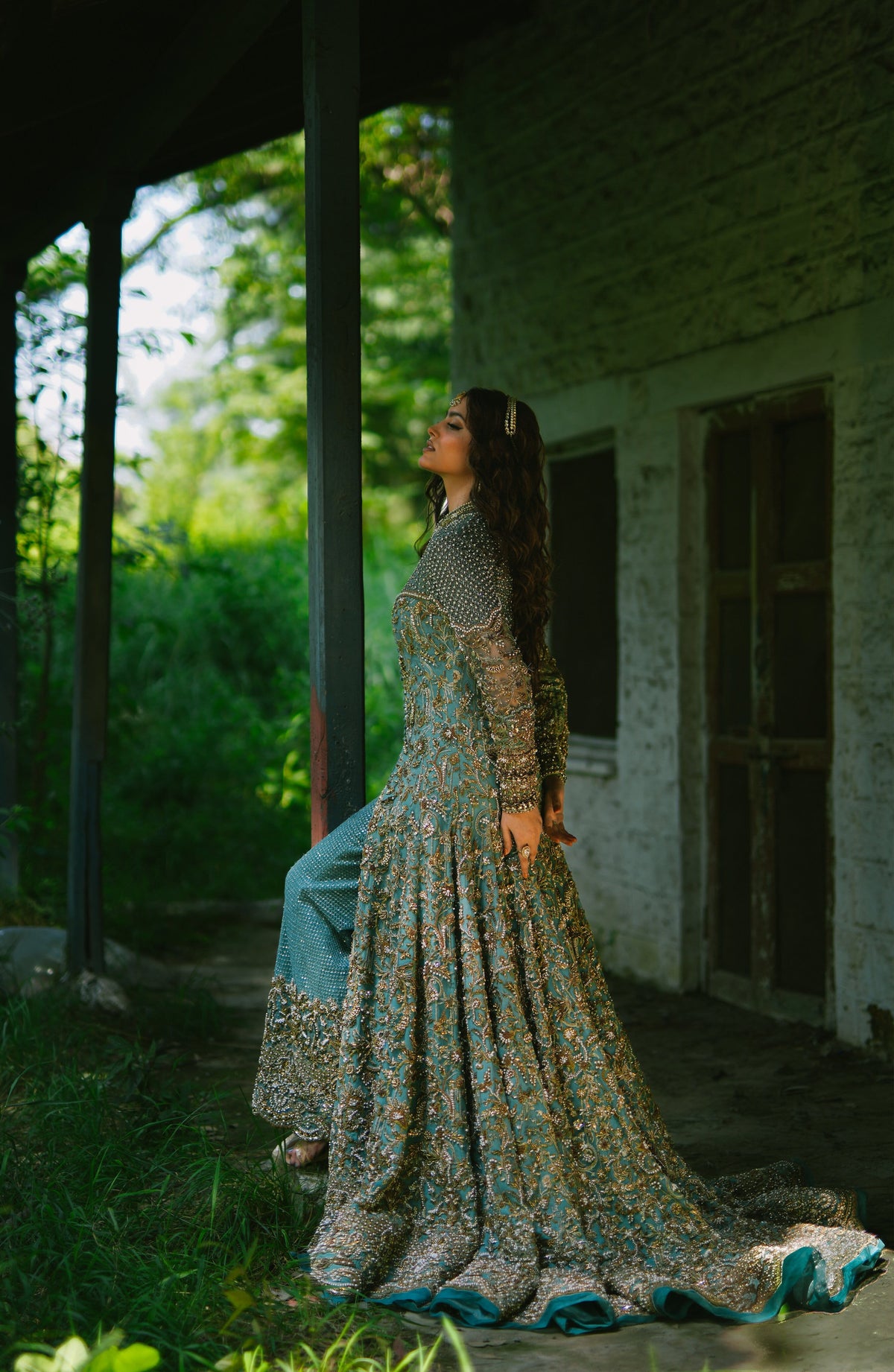 A woman wearing a long, heavily embroidered traditional gown with a teal and gold patterned skirt and a metallic silver sleeveless overlay, standing on a rustic porch with lush green foliage in the background.