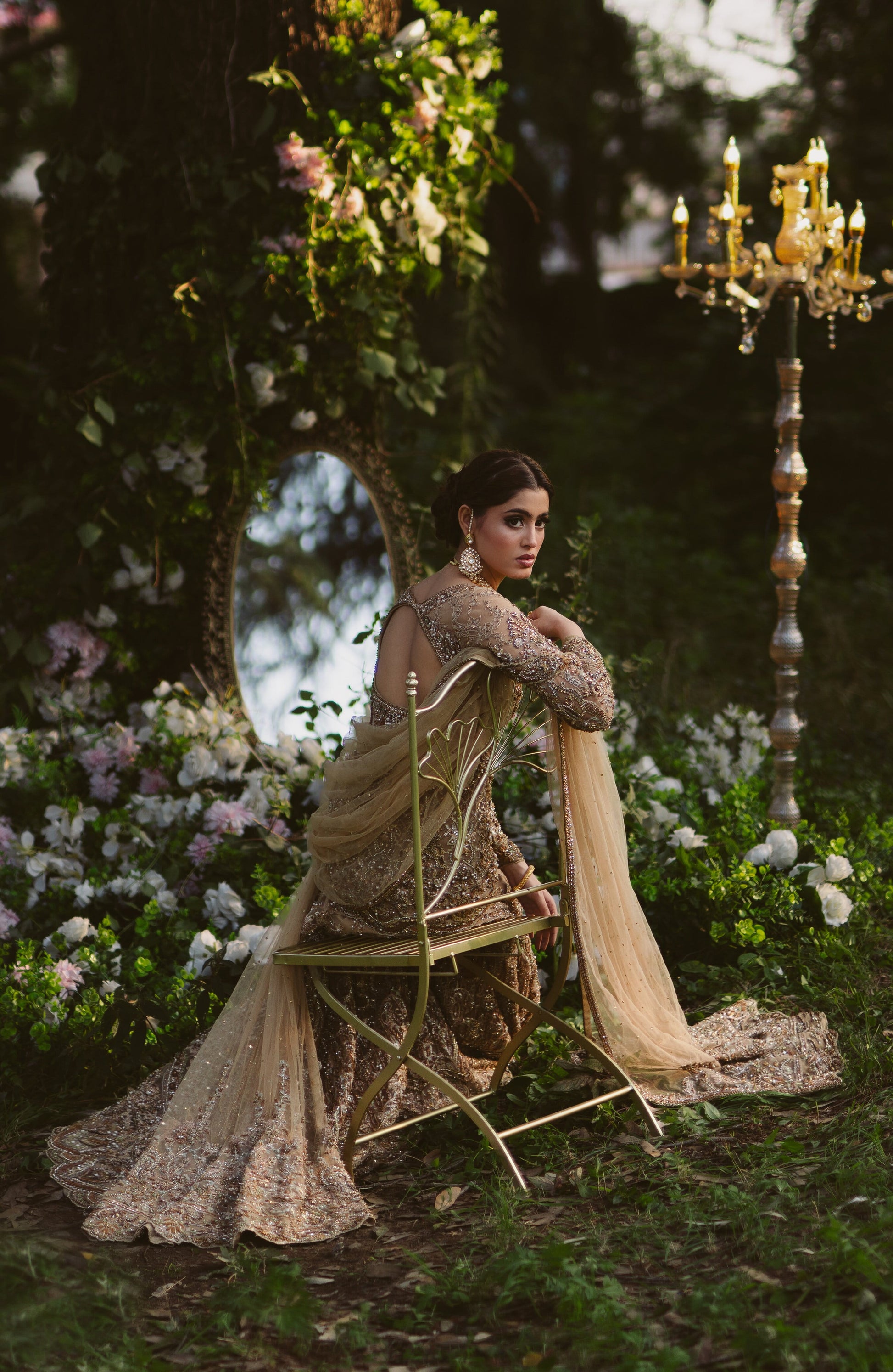 A woman in an ornate beige and gold embroidered gown sits on a gold folding chair in a garden setting with flowers, a mirror, and a tall candelabra.