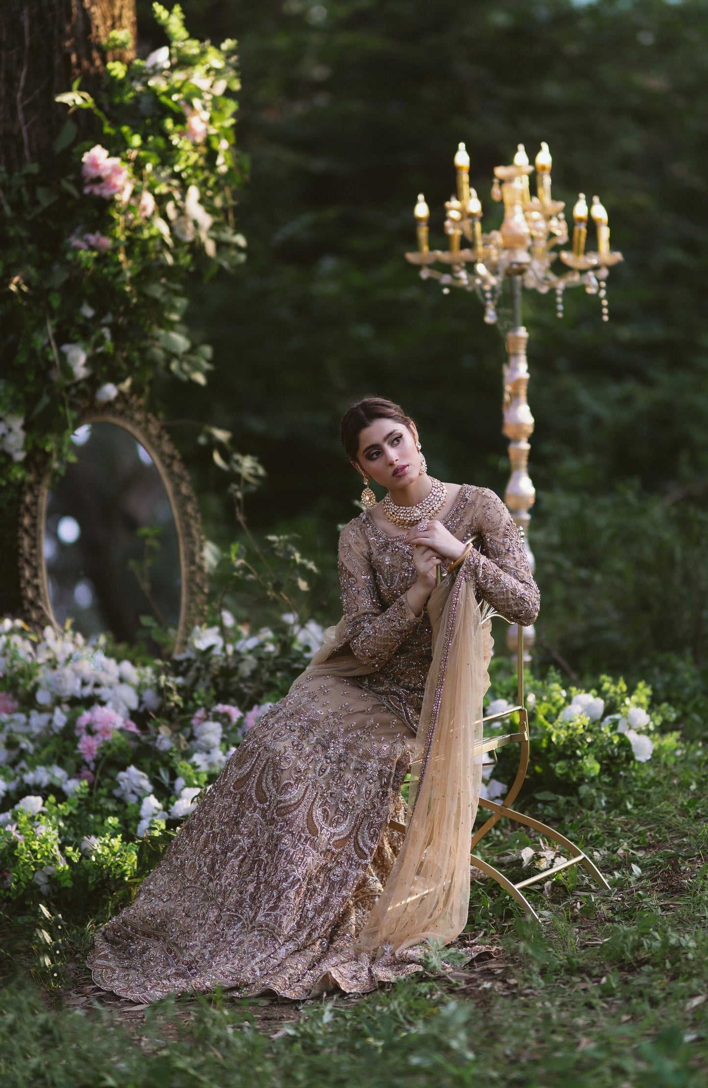A woman in an ornate beige and gold embroidered gown sits on a gold candelabra in a lush garden setting with flowers and greenery.