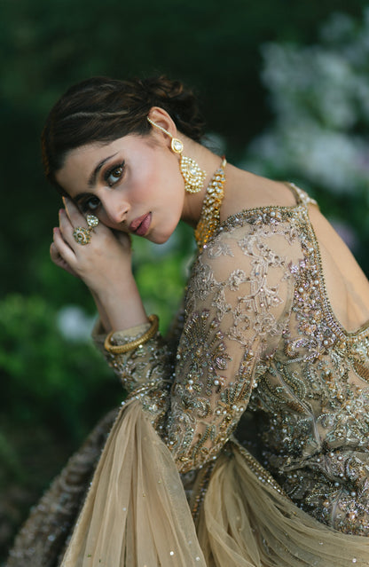 A woman in an ornate beige and gold embellished traditional outfit with heavy beadwork, posing outdoors against a blurred green background. She is accessorized with gold jewelry.