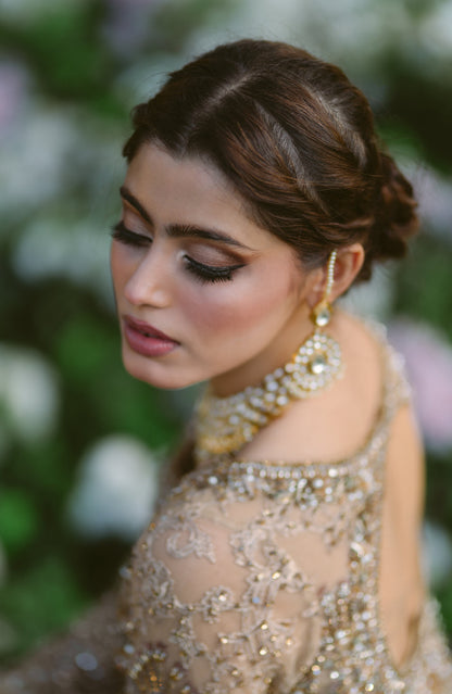 A woman in profile wearing an ornate beige embellished gown with a matching pearl and gold statement necklace and earring, set against a soft, blurred green and white garden background.