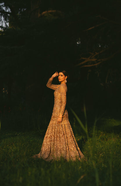 A woman stands in a grassy outdoor setting at dusk, wearing a long-sleeved, floor-length beige gown covered in intricate beadwork and sequins that catch the warm light.