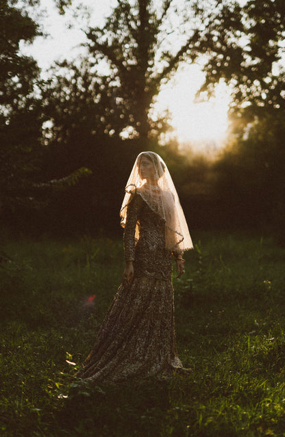 A woman in a long sheer lace dress and veil stands in a grassy outdoor area with trees and warm sunlight in the background.