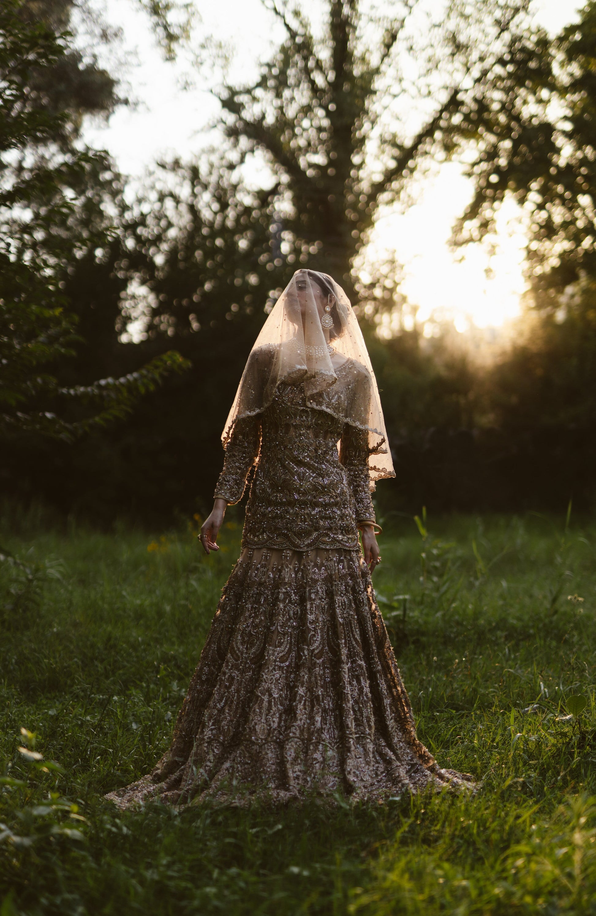 A woman wearing a long-sleeved, heavily embellished lace gown and a sheer veil stands in a grassy outdoor field at sunset, with trees in the background.
