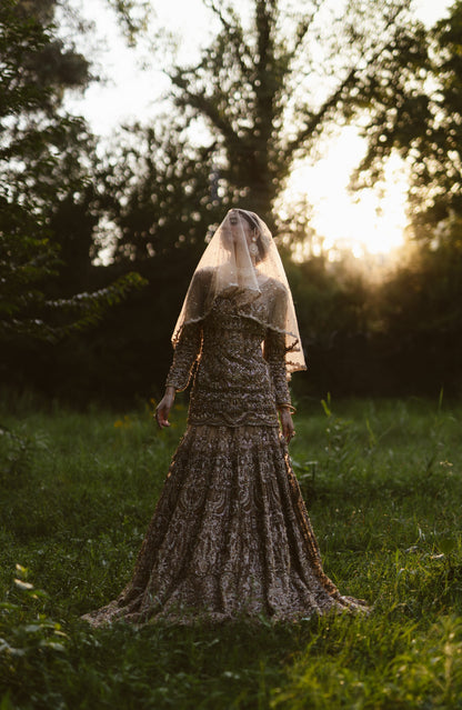 A woman wearing a long-sleeved, heavily embellished lace gown and a sheer veil stands in a grassy outdoor field at sunset, with trees in the background.