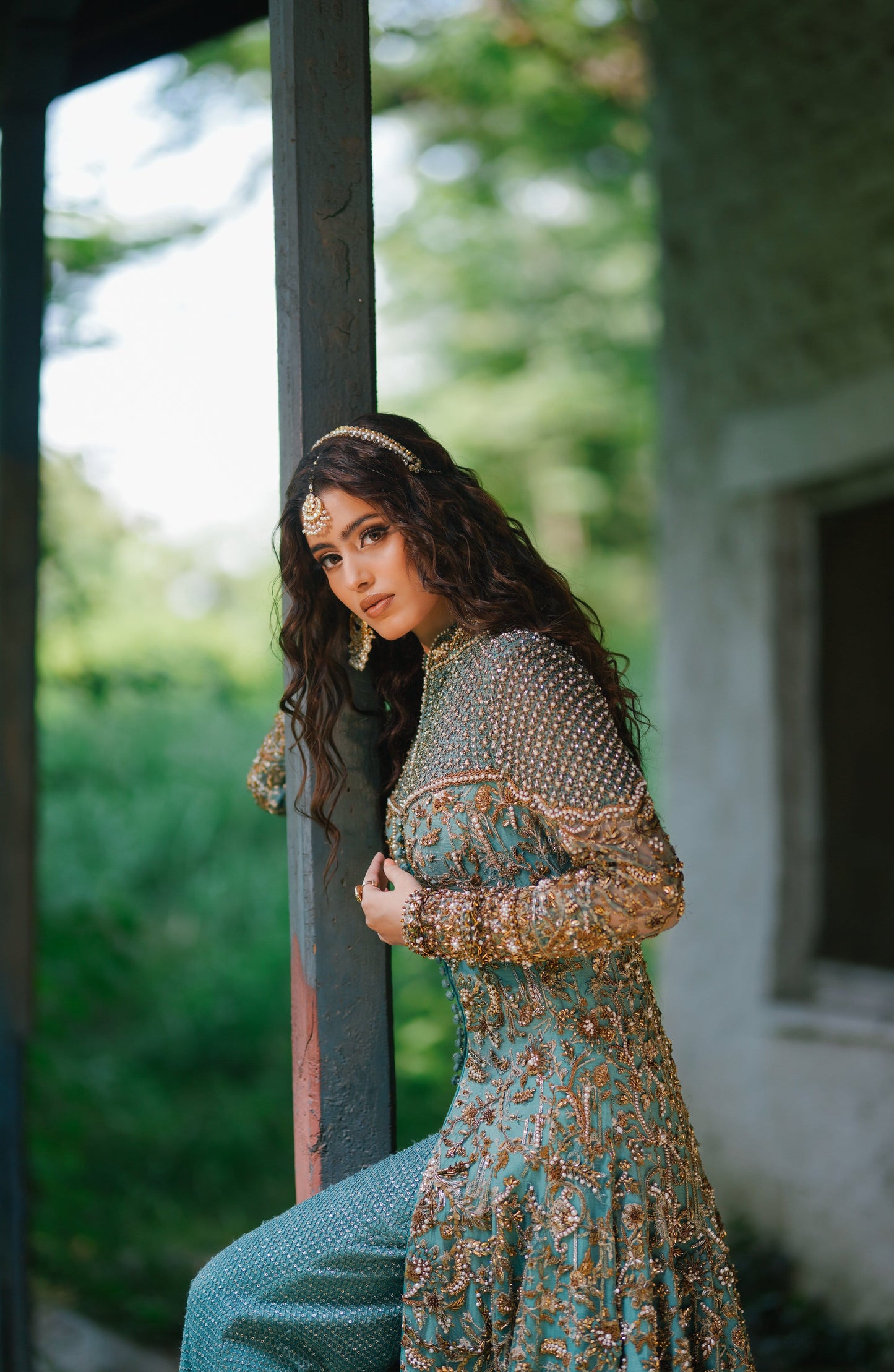 A woman wearing an ornate teal and gold embellished traditional dress leans against a dark wooden post in an outdoor setting with blurred green foliage and a stone wall in the background.