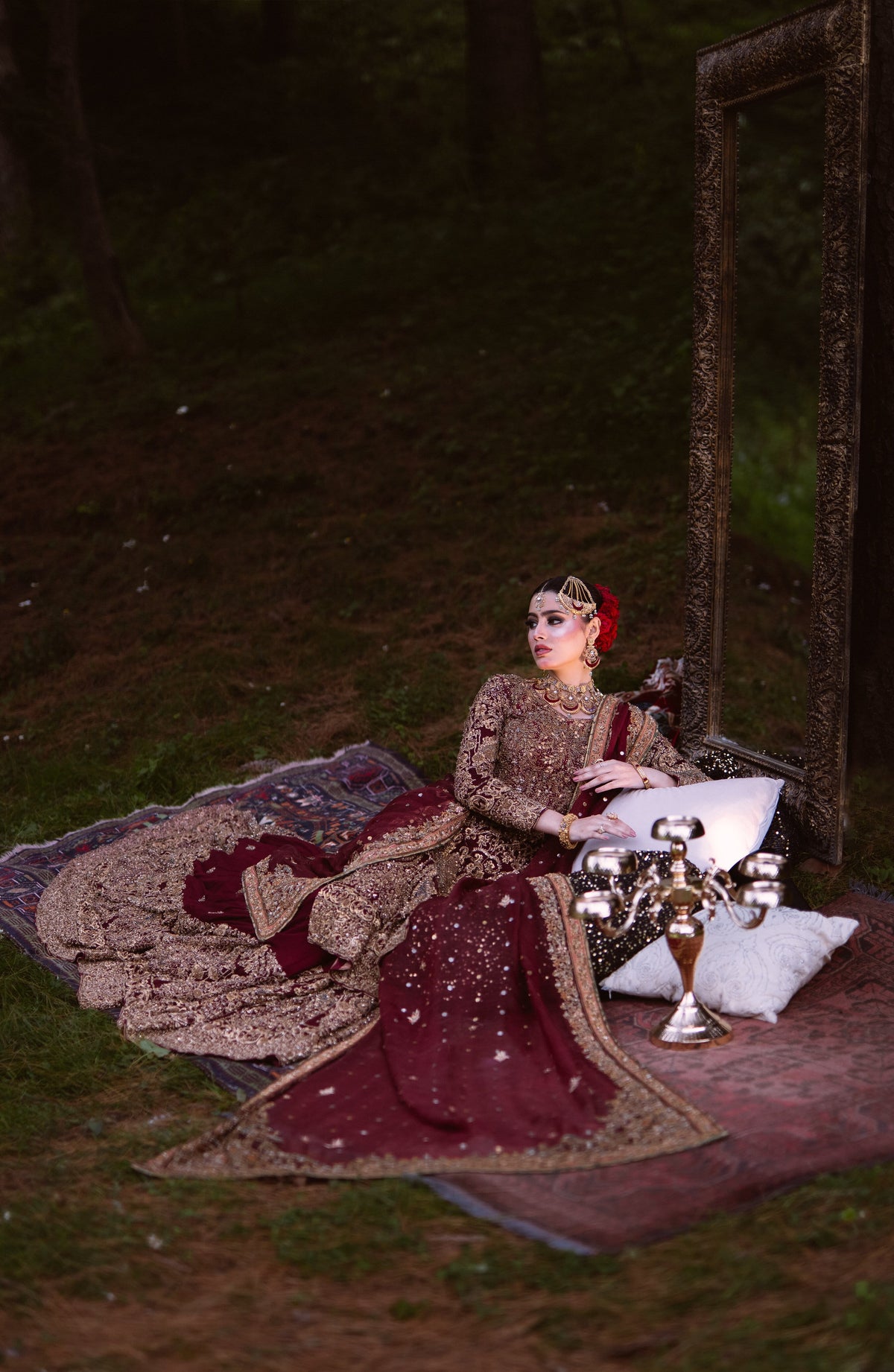 A woman in an ornate traditional maroon and gold embroidered dress reclines on a rug beside a large mirror in a dark outdoor setting.