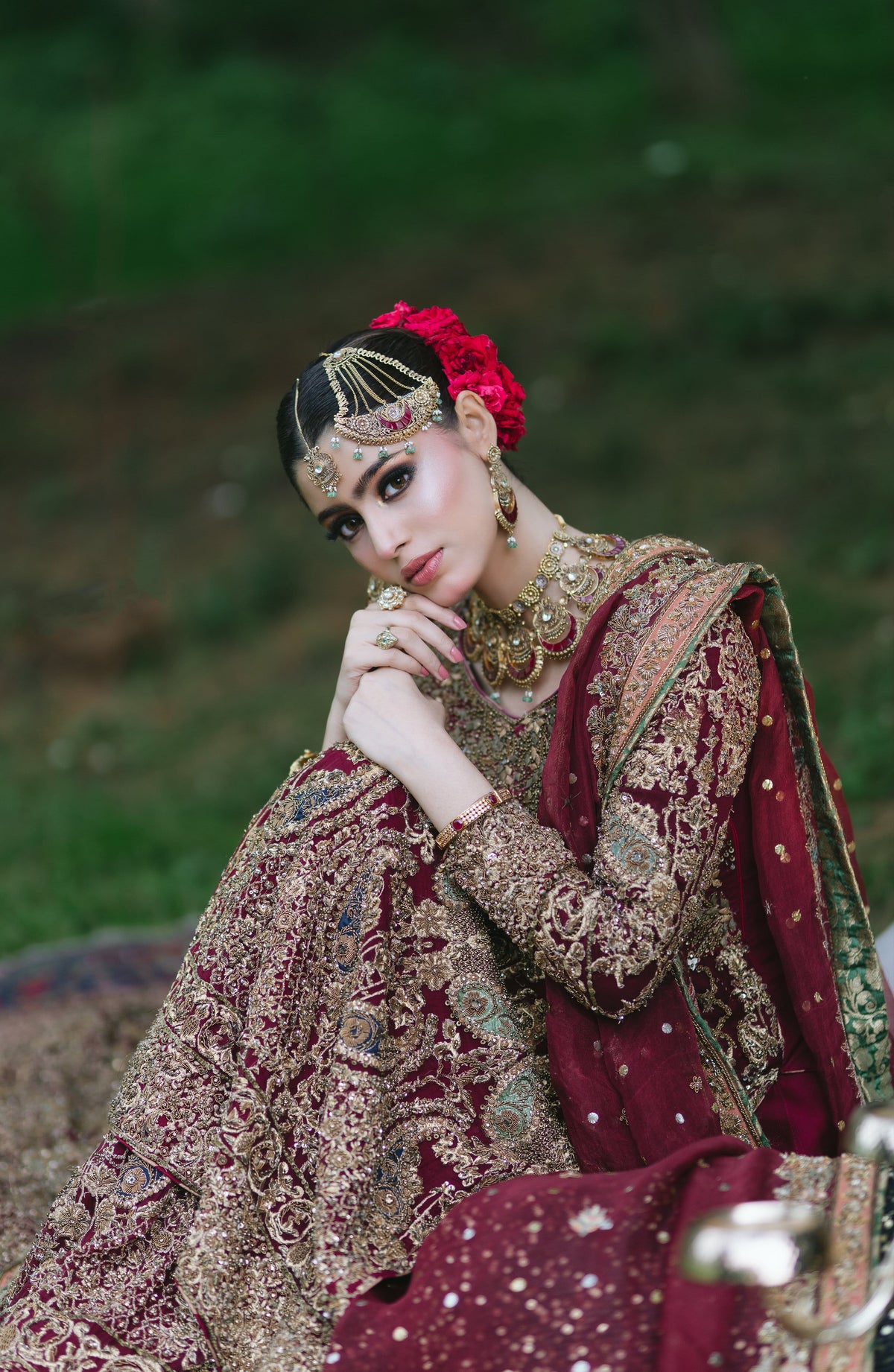 A woman wearing an ornate maroon and gold embroidered traditional outfit with a red floral headpiece, posed outdoors against a blurred green background.