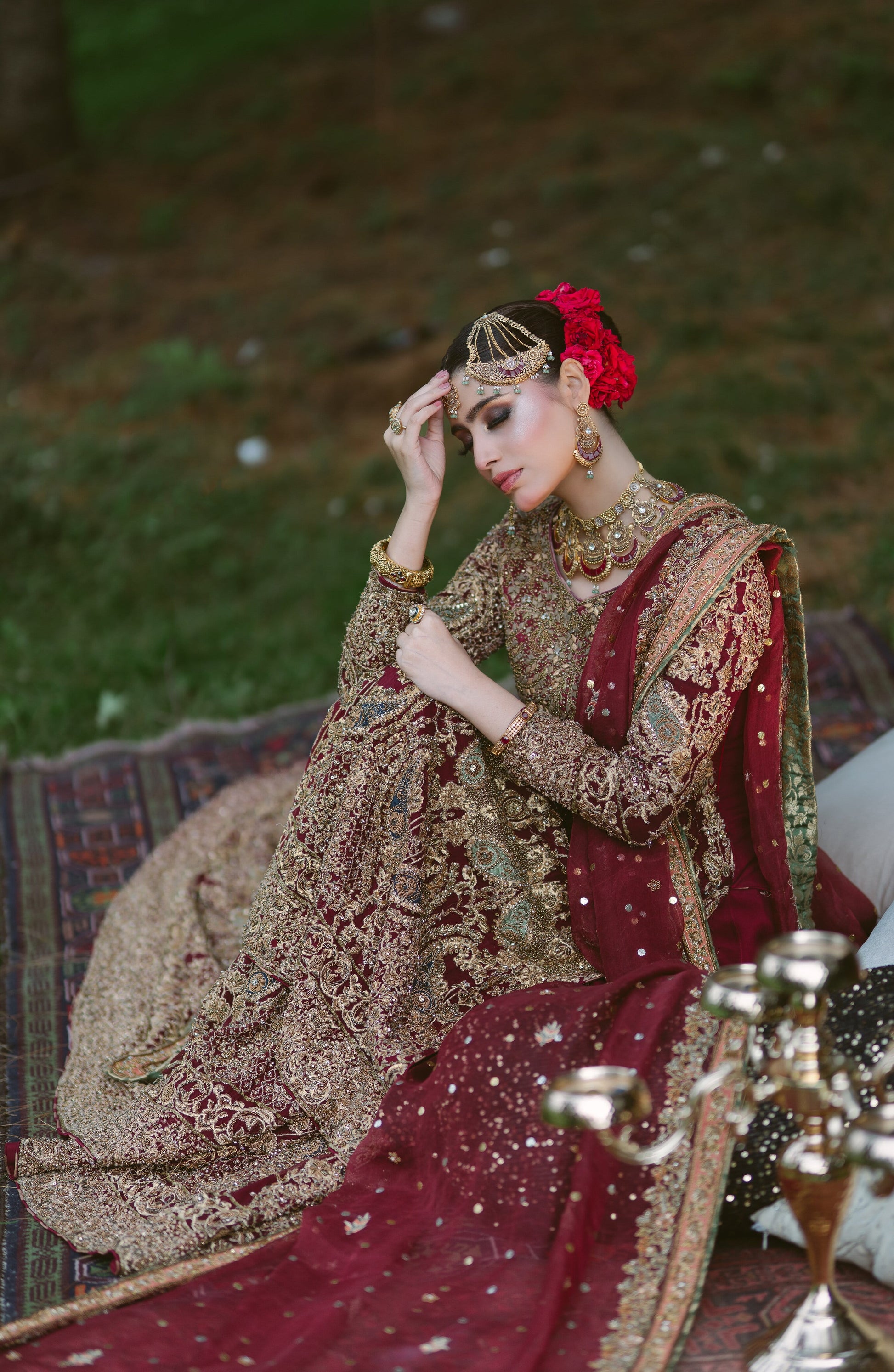 A woman wearing an ornate maroon and gold embroidered traditional outfit with a red floral hair accessory, seated outdoors on a patterned rug with decorative metal vessels in the foreground.