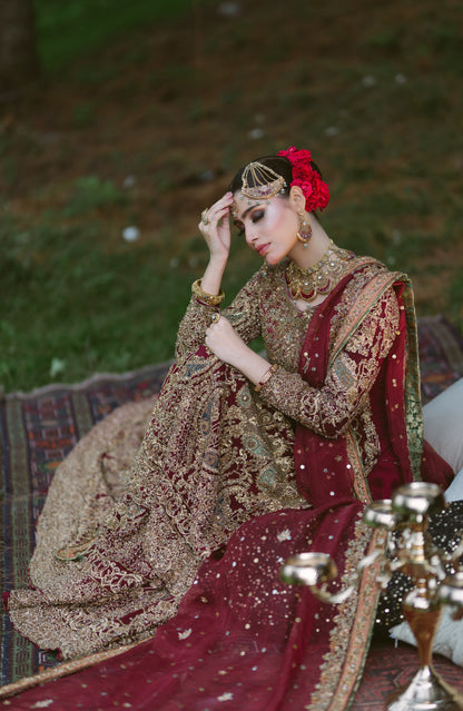 A woman wearing an ornate maroon and gold embroidered traditional outfit with a red floral hair accessory, seated outdoors on a patterned rug with decorative metal vessels in the foreground.