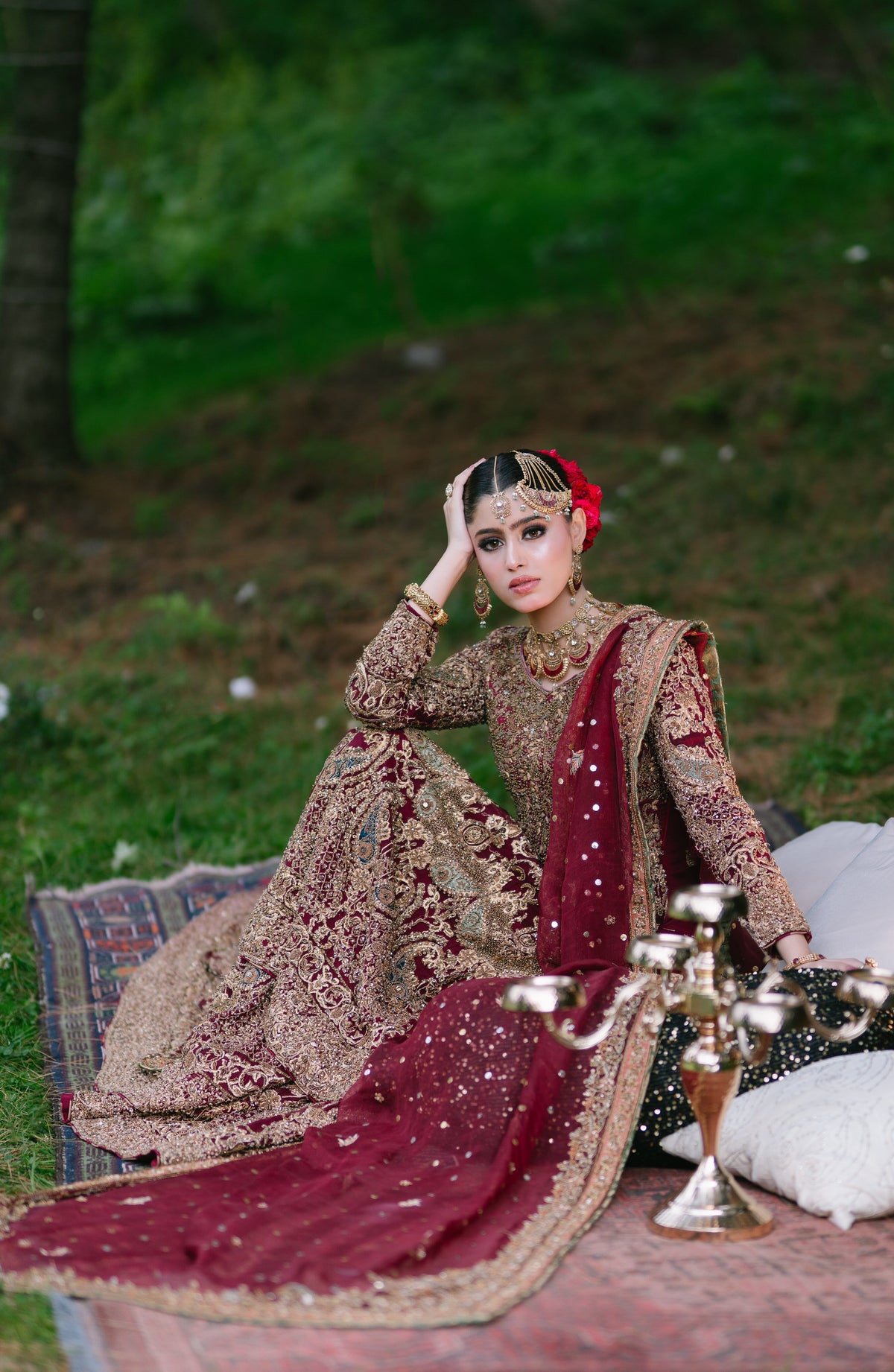 A woman wearing an ornate traditional bridal outfit sits outdoors on a rug with cushions. The outfit is heavily embroidered in beige and gold with a deep maroon dupatta and red floral hair accessory.