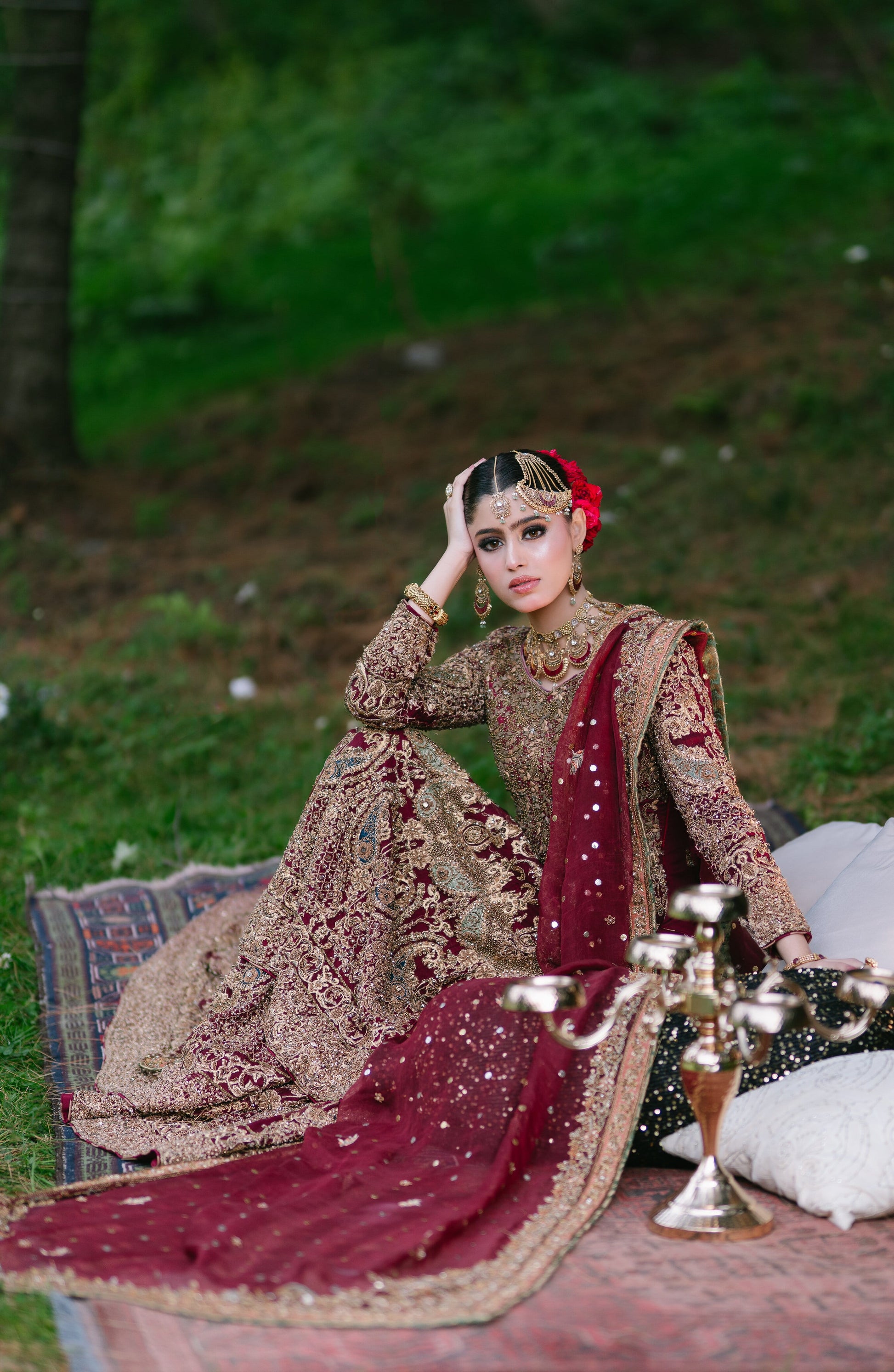 A woman wearing an ornate traditional bridal outfit sits outdoors on a rug with cushions. The outfit is heavily embroidered in beige and gold with a deep maroon dupatta and red floral hair accessory.