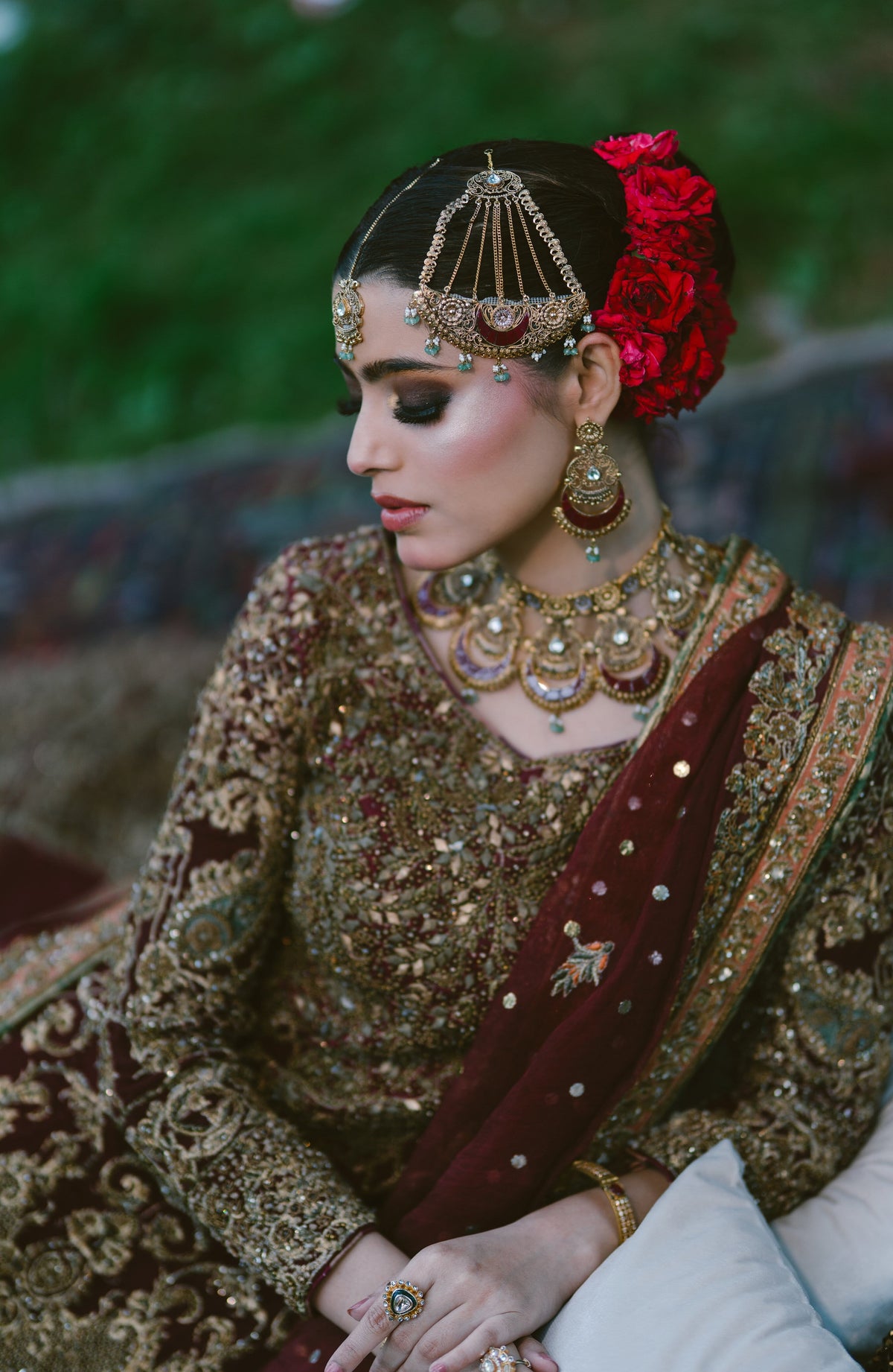A woman wearing traditional South Asian bridal attire, featuring an intricately embroidered brown and gold lehenga, a deep maroon dupatta, a red floral hair accessory, and elaborate gold jewelry, captured in a soft-focus outdoor setting.