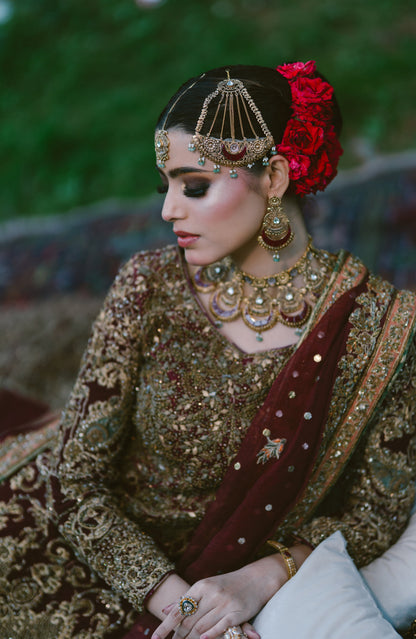 A woman wearing traditional South Asian bridal attire, featuring an intricately embroidered brown and gold lehenga, a deep maroon dupatta, a red floral hair accessory, and elaborate gold jewelry, captured in a soft-focus outdoor setting.