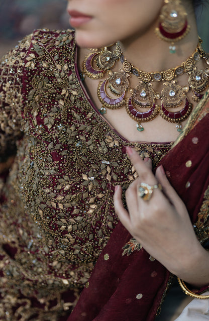 Close-up of a person wearing an ornate maroon and gold embroidered traditional outfit with a matching draped scarf, paired with a large gold statement necklace and earrings.