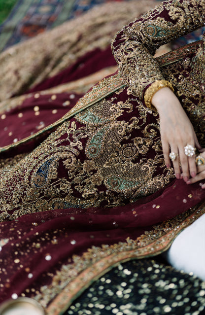 A close-up of a person wearing an ornate traditional outfit with a deep maroon velvet jacket heavily embellished with gold embroidery, paired with a matching maroon dupatta and a black sequined garment.