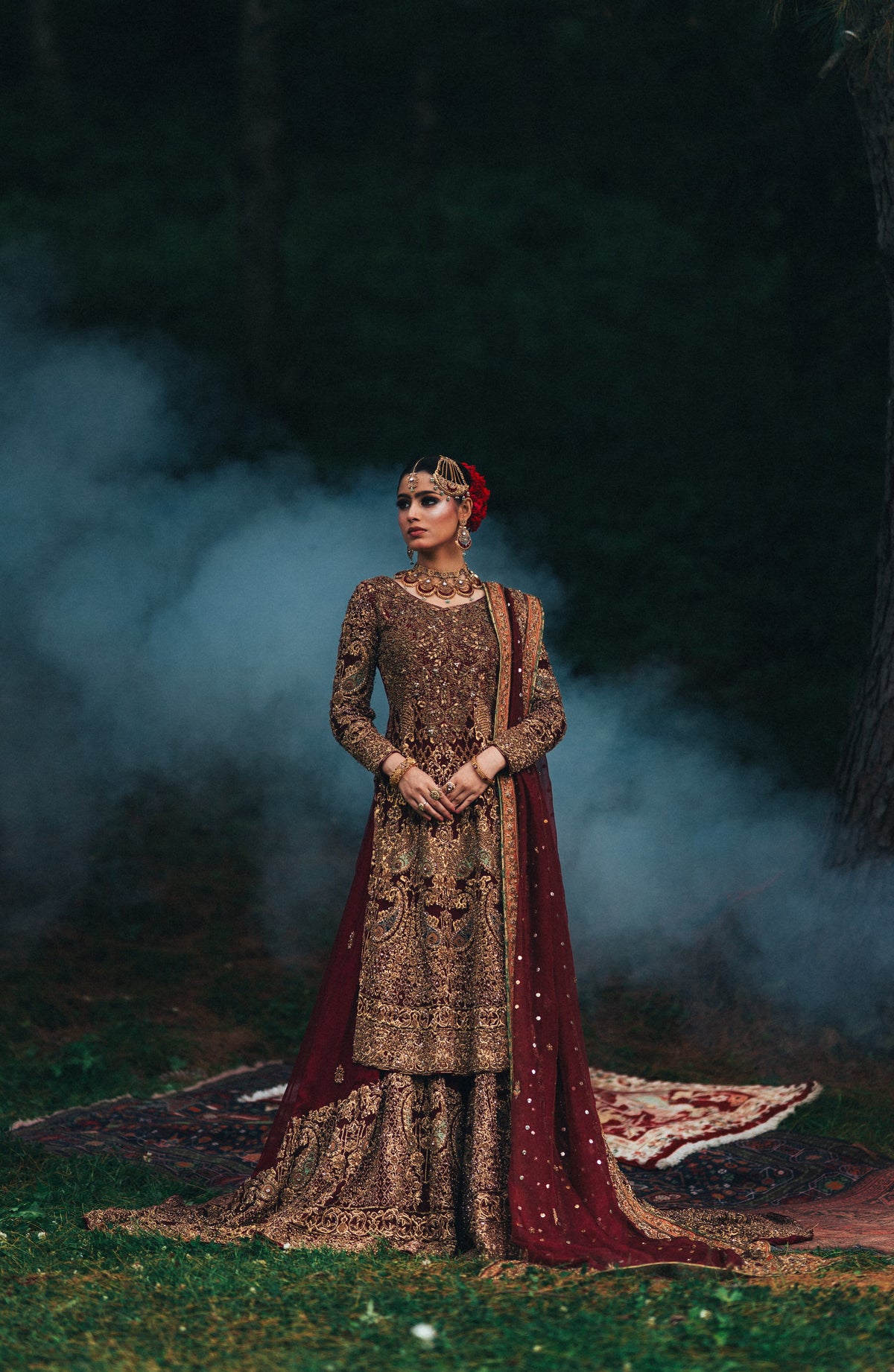 A woman stands outdoors in a forest clearing wearing an ornate traditional outfit with heavy embroidery. The garment is primarily deep brown with intricate gold detailing, paired with a draped maroon dupatta and a red floral headpiece.