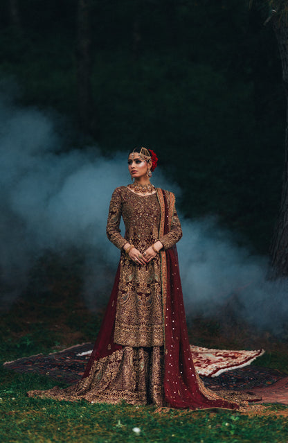A woman stands outdoors in a forest clearing wearing an ornate traditional outfit with heavy embroidery. The garment is primarily deep brown with intricate gold detailing, paired with a draped maroon dupatta and a red floral headpiece.