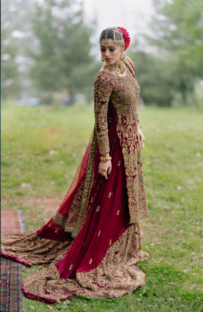 A woman wearing an ornate traditional embroidered gown stands outdoors on grass. The dress is deep maroon with extensive gold embroidery and a matching sheer dupatta, while she wears a red floral headpiece and heavy gold jewelry.