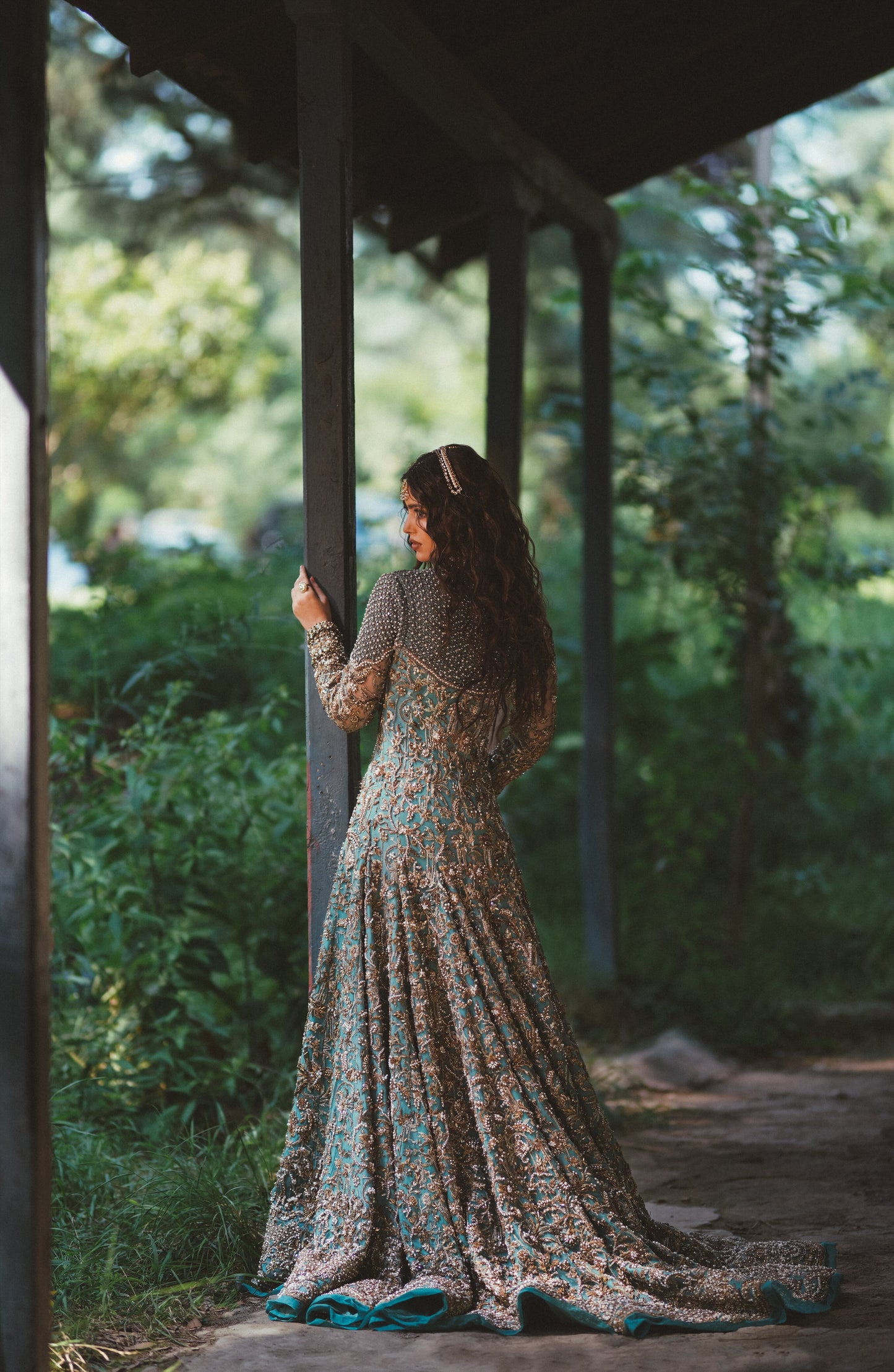 A woman stands on a covered walkway outdoors, wearing a long-sleeved floor-length gown with a dense floral and paisley print in beige, brown, and teal. She is seen from behind, looking out toward a lush green garden.