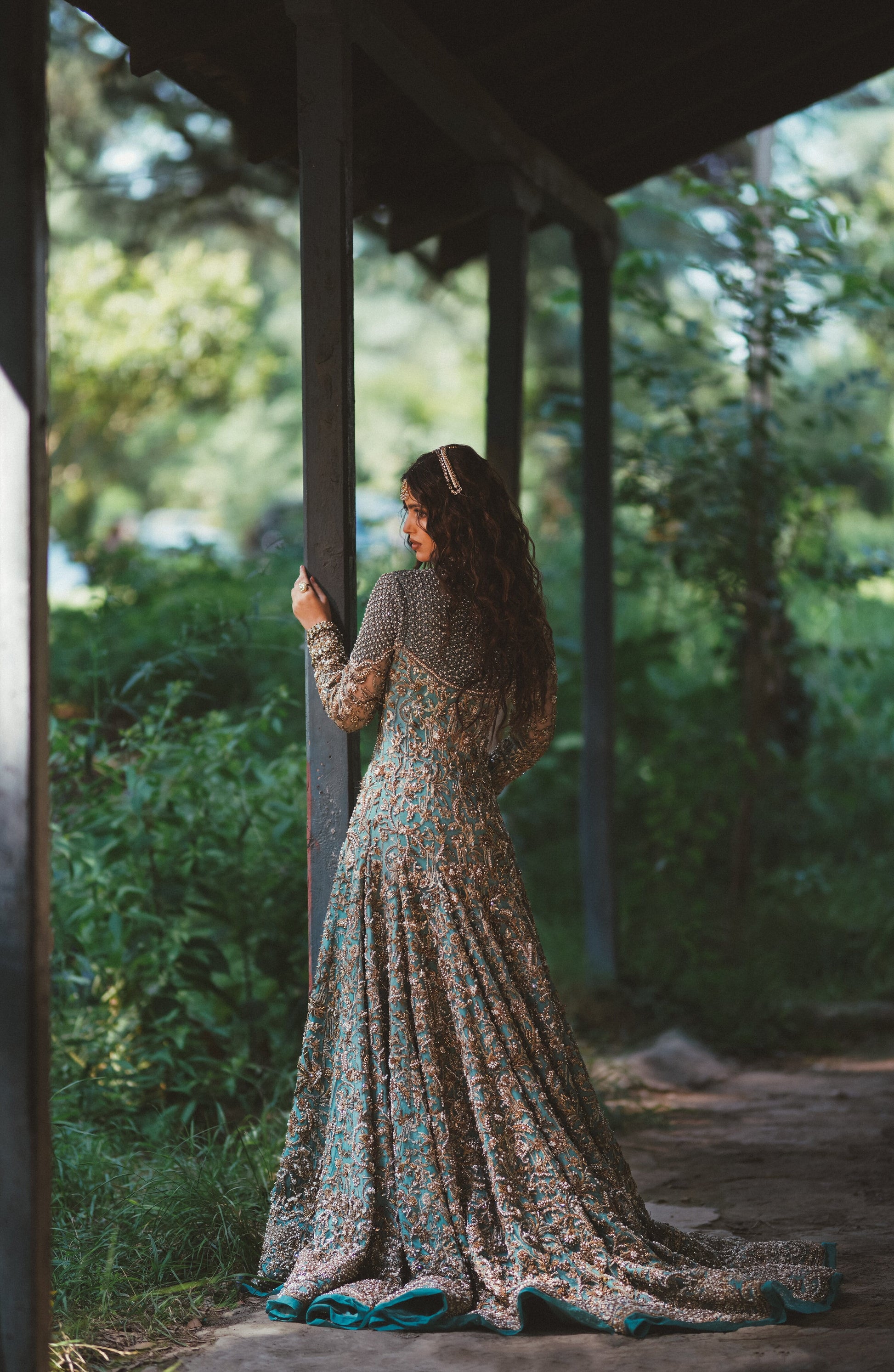 A woman stands on a covered walkway outdoors, wearing a long-sleeved floor-length gown with a dense floral and paisley print in beige, brown, and teal. She is seen from behind, looking out toward a lush green garden.
