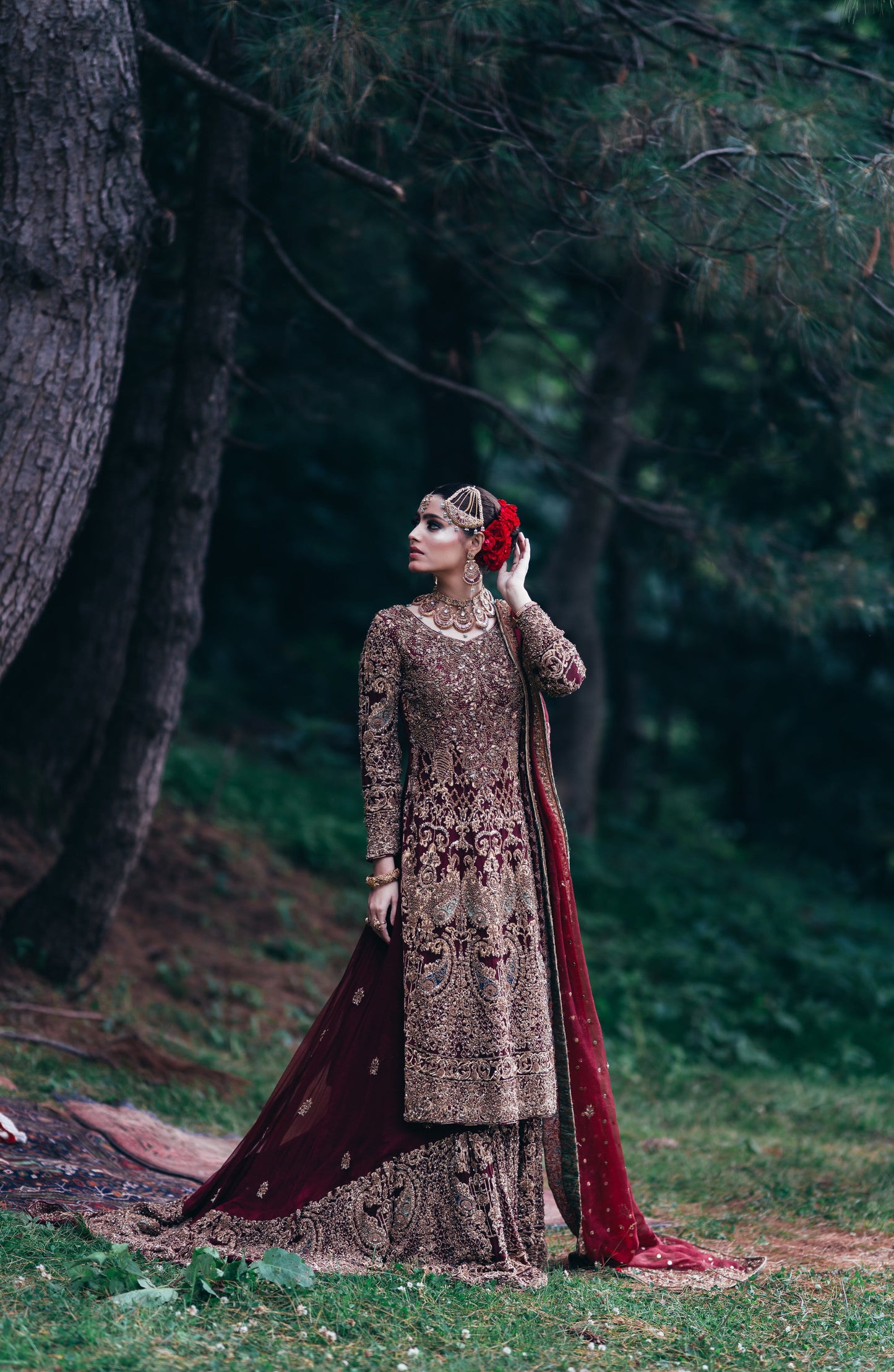 A woman stands outdoors in a wooded setting wearing an ornate traditional outfit with a heavily embroidered brown and gold kurta, a matching skirt, and a deep red dupatta draped over the ground. She is holding a red flower to her hair.