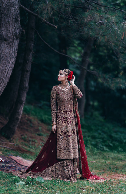 A woman stands outdoors in a wooded setting wearing an ornate traditional outfit with a heavily embroidered brown and gold kurta, a matching skirt, and a deep red dupatta draped over the ground. She is holding a red flower to her hair.