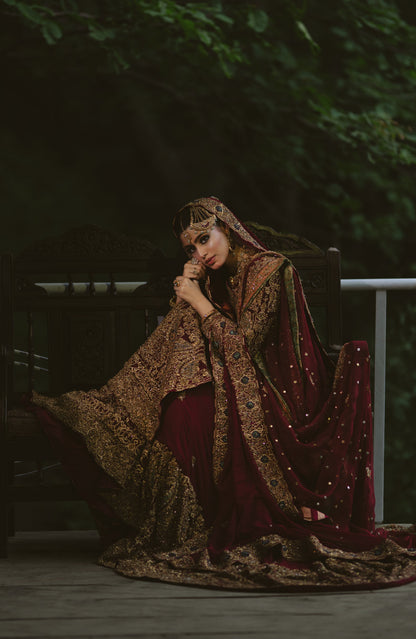 A woman in an ornate deep maroon and gold embroidered traditional outfit with a matching veil, seated on a dark wooden bench in a dim outdoor setting with green foliage in the background.