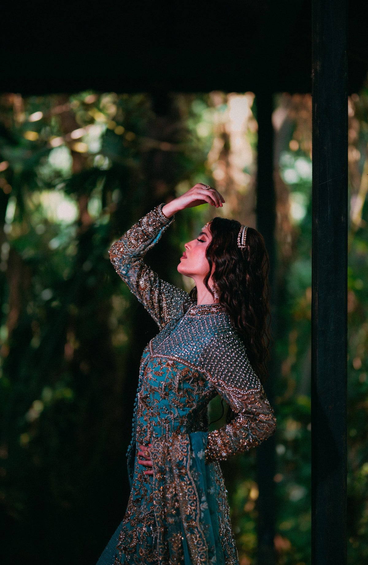 A woman wearing a long-sleeved teal and gold heavily embellished traditional dress, posing in a dark, rustic wooden structure with a lush green forest background.