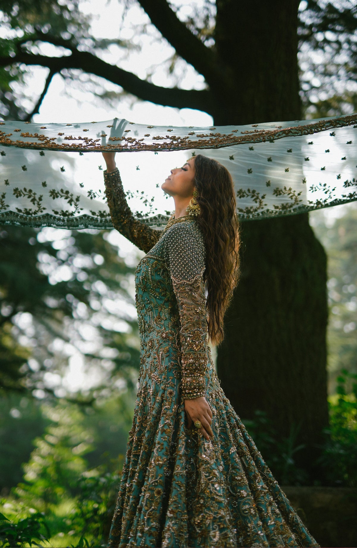 A woman in a long, heavily embroidered teal and gold gown stands outdoors under a large tree, holding up a sheer white embroidered scarf.