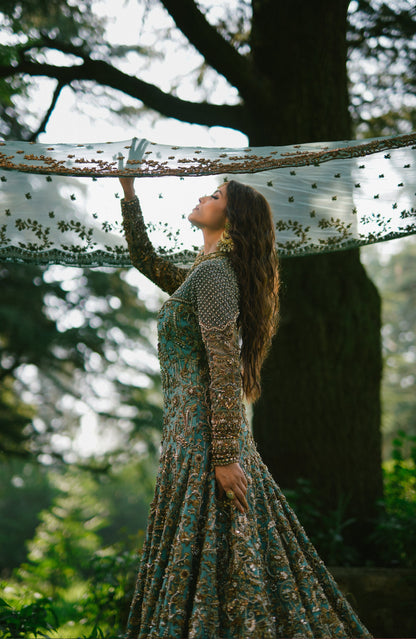A woman in a long, heavily embroidered teal and gold gown stands outdoors under a large tree, holding up a sheer white embroidered scarf.