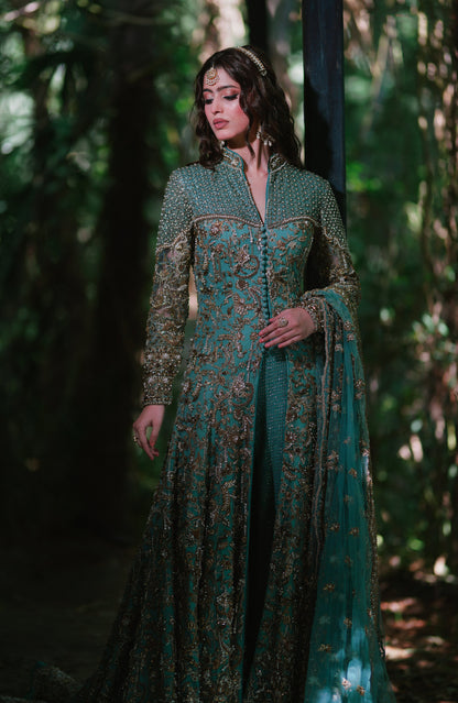 A woman wearing an ornate teal green embroidered traditional gown with gold detailing, standing in a dark, lush forest setting.