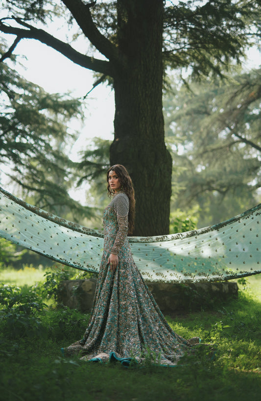 A woman stands in a sun-dappled forest wearing a long, flowing floral gown with sheer sleeves, with a sheer patterned fabric draped across a tree trunk behind her.