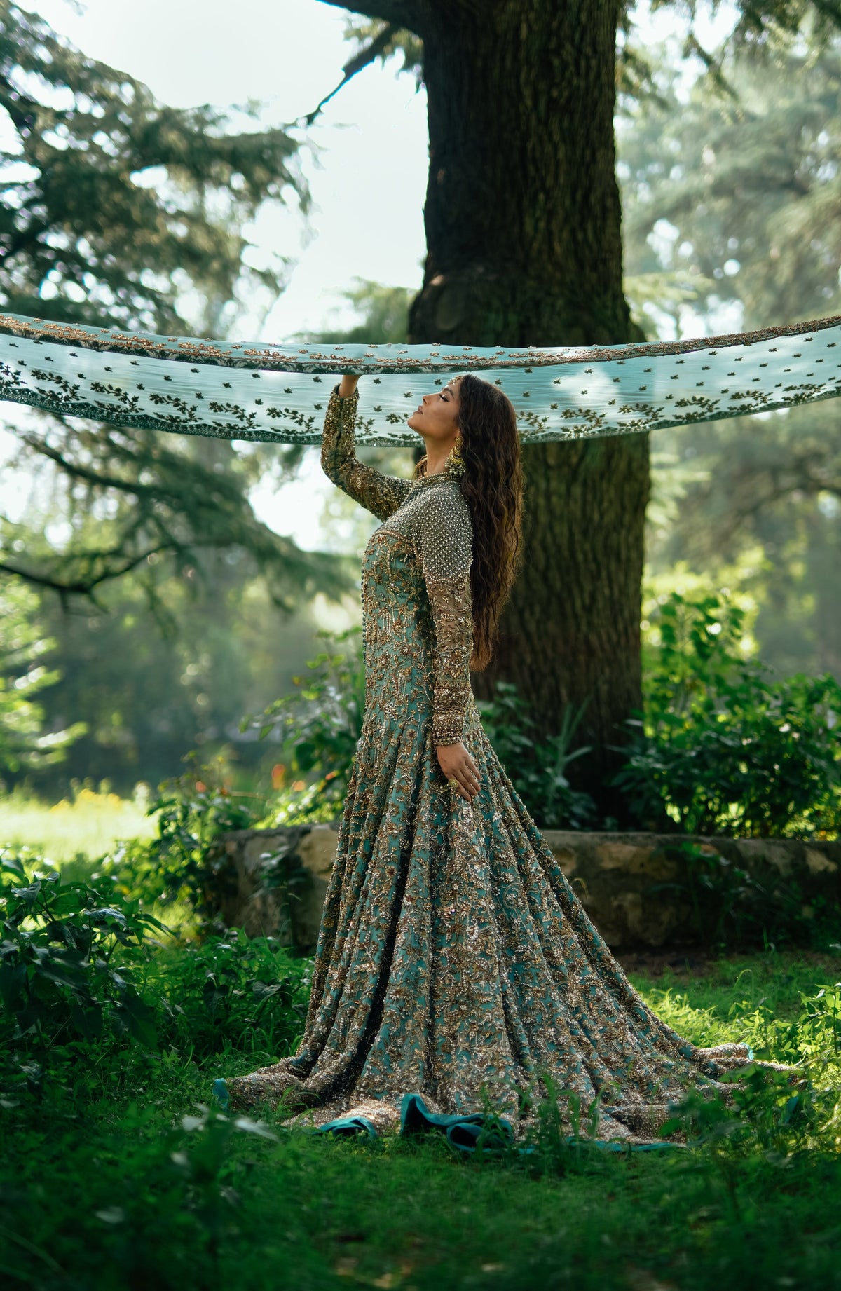 A woman in a long, flowing patterned dress stands outdoors in a sunlit garden, holding up a sheer matching fabric draped across trees.