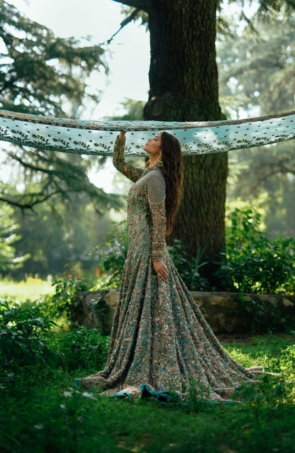 A woman in a long, flowing patterned dress stands outdoors in a sunlit garden, holding up a sheer matching fabric draped across trees.