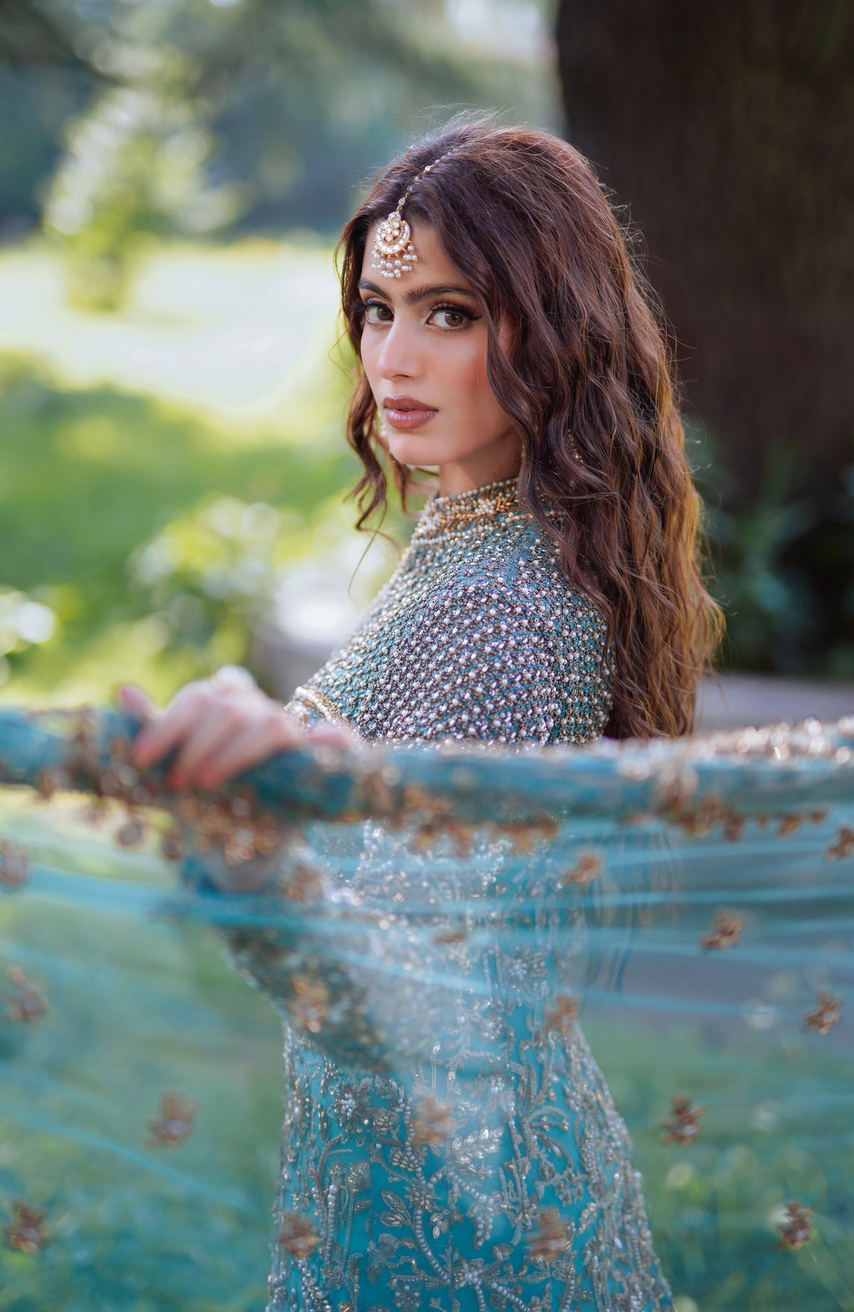 A woman outdoors wearing a heavily embellished light blue traditional outfit with gold embroidery and holding a sheer blue dupatta with gold motifs.