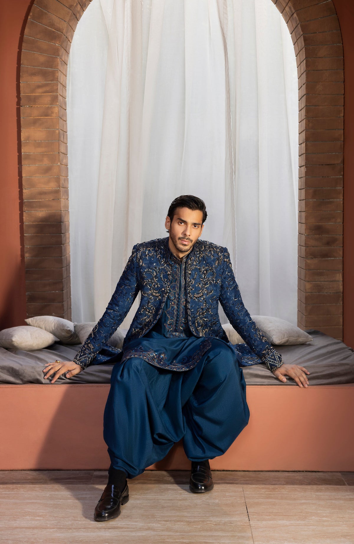 A man wearing a traditional Indian outfit with a navy blue embroidered sherwani and matching harem pants, sitting on a cushioned bench in front of a white curtain and brick archway.
