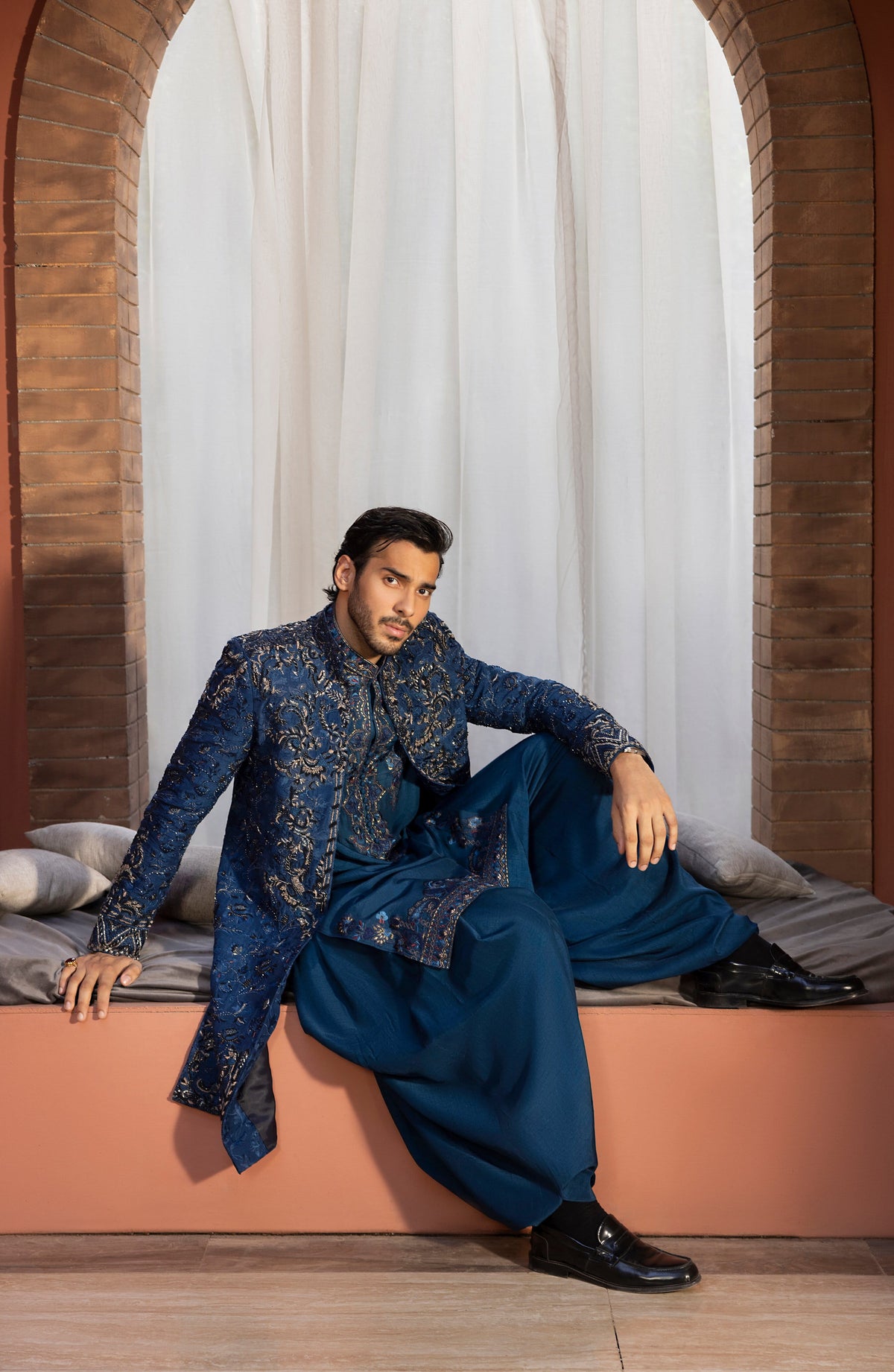 A man models a traditional Indian outfit consisting of a navy blue embroidered sherwani jacket and matching wide-leg trousers, seated on a terracotta-colored platform in front of white curtains and brick arches.