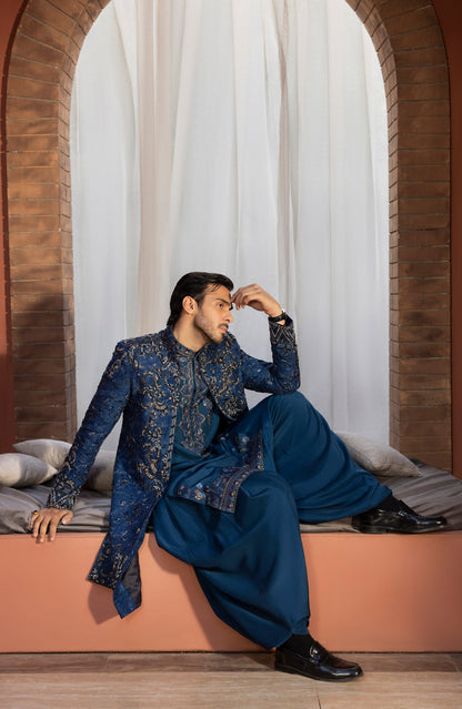 A man wearing a traditional Indian outfit with a navy blue and gold embroidered sherwani jacket and matching wide-leg trousers, sitting on a cushioned bench in front of a white draped window setting with brick arches.