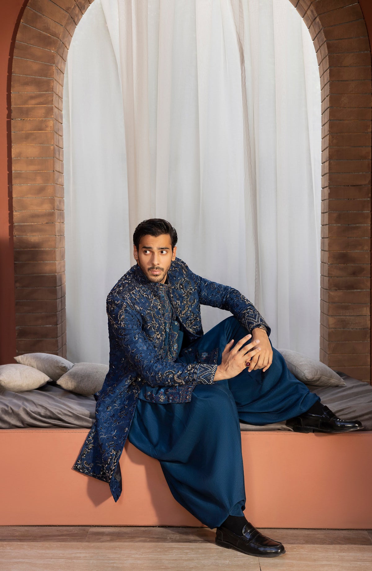A man wearing a traditional Indian outfit consisting of a navy blue embroidered kurta and matching trousers, sitting on a terracotta-colored window seat with white curtains and brick arches in the background.